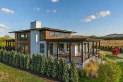 Modern two-story home with stone and wood siding, large windows, and a wraparound porch set against a scenic landscape.
