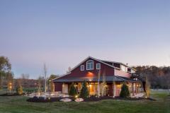 Exterior view of the red party barn with expansive yard and welcoming landscaping. The glow of the landscape lighting gives the illusion that it is sparkling.