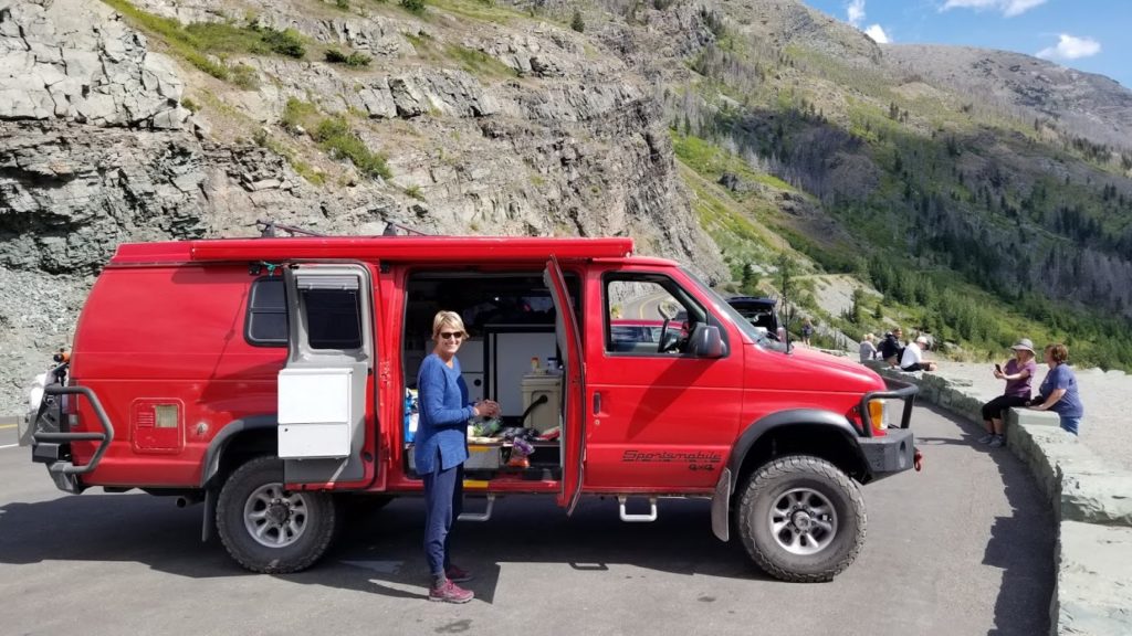 A woman standing with a 2001 Ford E350 extended body Sportsmobile with penthouse pop-top and a Quigley 4X4 conversion.