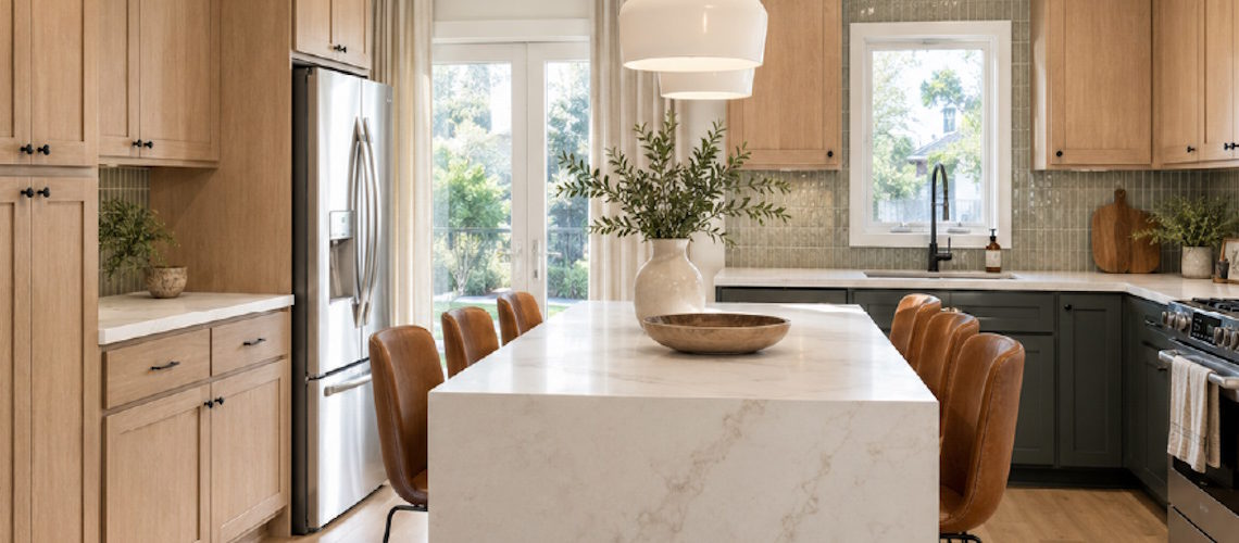 A bright, modern kitchen featuring light oak cabinetry, dark green lower cabinets, a marble waterfall island with leather barstools.
