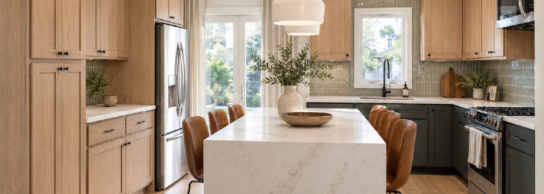 A bright, modern kitchen featuring light oak cabinetry, dark green lower cabinets, a marble waterfall island with leather barstools.