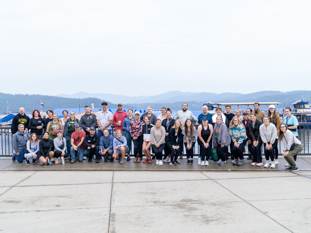 A large group ready to go on a hike, standing in front of lake Coeur d'Alene. 